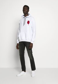 White hoodie with a red heart graphic, black drawstrings, and front pocket, paired with dark jeans and white sneakers.