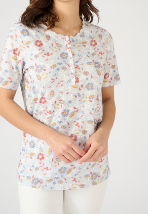 Femme portant une chemise à manches courtes à imprimé floral avec des boutons et un col festonné, debout les mains croisées devant elle.