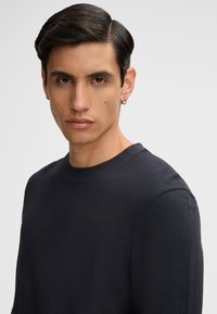 Zwarte langwerpige shirt met een gestructureerde stof, ronde halsontwerp en zichtbare stiksels bij de halslijn. Model toont een neutrale uitstraling.