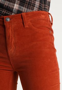 Pantalon en velours côtelé couleur rouille porté avec une chemise à carreaux marron et noire rentrée, montrant les détails des boutons et des passants de ceinture.