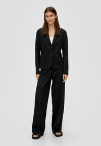 Vrouw die tegen een effen achtergrond staat en een zwarte blazer, wijde broek, witte top eronder en zwarte sandalen draagt.