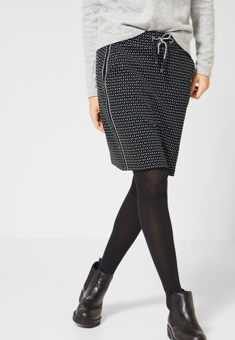 Femme portant une jupe noire à motifs longueur genou avec cordon, collants noirs, bottines noires et un pull gris clair.