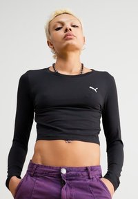 Top cropped preto de manga longa com um logo branco da Puma no lado esquerdo do peito; calças roxas com detalhe de botão frontal. Textura suave e ajustada.
