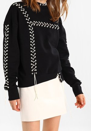 Svart sweatshirt med vit snörning, med rund halsringning, långa ärmar och dekorativa dragsnören vid nederkant. Vit kjol under.