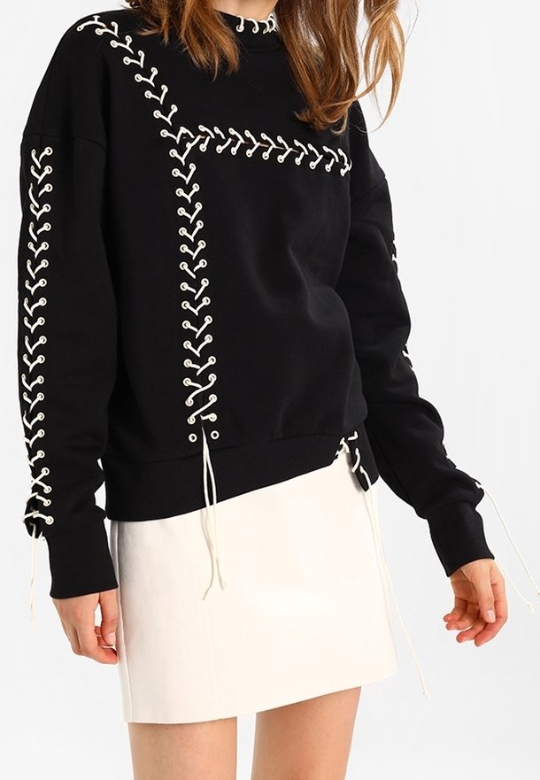 Musta huppari, jossa on valkoista nauhaketjua, pyöreä kaula-aukko, pitkät hihat ja koristeelliset nyörit helman kohdalla. Valkoinen hame sen alla.