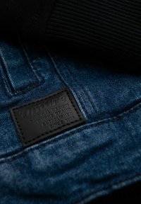 Tessuto di denim con un lavaggio blu scuro, caratterizzato da una toppa in pelle nera embossata con la scritta "Ombre" e dettagli del marchio. Cuciture testurizzate visibili.