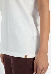 Hvid bomulds T-shirt med korte ærmer, lige snit, sideslids og et lille brun lædermærke ved kanten. Bæres med mørk burgunder bukser.