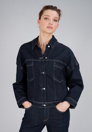 Frau mit hellem Teint und zurückgebundenen Haaren, die eine dunkelblaue Jeansjacke und Jeans trägt, steht mit den Händen in den Taschen vor einem hellgrauen Hintergrund.