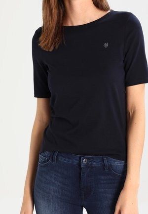 Frau trägt ein schlichtes schwarzes T-Shirt mit kurzen Ärmeln und einem kleinen Logo auf der Brust sowie dunkelblaue Jeans.