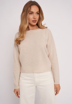 Kobieta o falowanych blond włosach ubrana w beżowy sweter z długim rękawem i białe spodnie, stojąca na tle jednolitego jasnego tła.