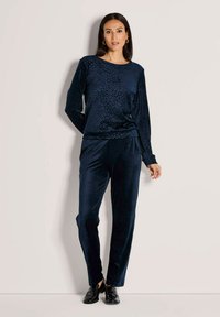 Vrouw staat tegen een beige muur en draagt een marineblauwe fluwelen top met panterprint en bijpassende broek met zwarte instappers.