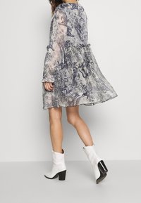 Průhledné květinové šaty v šedé a černé barvě, s volánkovými rukávy a splývavým lemem. Doplnené bílými kovbojskými botami s černým podpatkem.