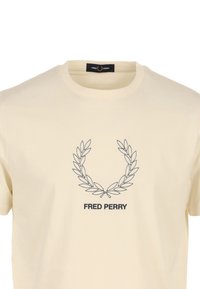 Camiseta de algodón color crema con una corona de laurel negra bordada y el texto "FRED PERRY" debajo. Diseño clásico de cuello redondo.
