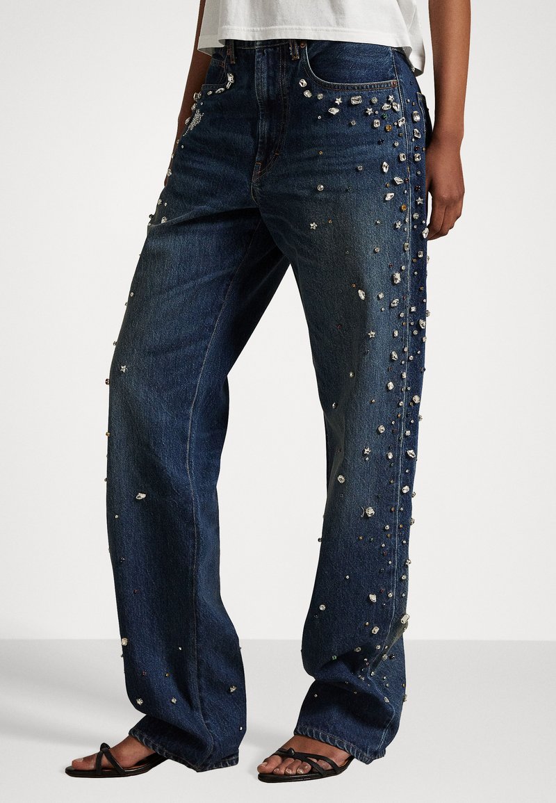 Dunkelblaue Jeans mit weitem Bein, verziert mit verschiedenen metallischen, sternförmigen und geometrischen Verzierungen entlang der Seiten.