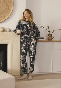 Melnas ar ziedu rakstu rakstīta mājas apģērbu komplekts ar garām piedurknēm, brīvu piegriezumu un aprocētiem biksu galiem. Istabas dekors ietver bēša vāzi ar ziediem.