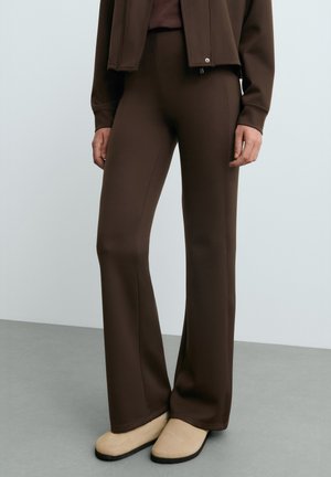 Persona che indossa pantaloni aderenti marrone scuro con una leggera svasatura e scarpe basse beige, in piedi su un pavimento grigio contro una parete chiara.