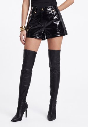 Vrouw die glanzende zwarte leren korte broek draagt, zwarte bovenbeen hoge stiletto laarzen en een gouden armband, staand tegen een witte achtergrond.