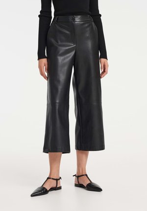Culottes en cuir noir avec taille haute, devant plat et jambes larges. Texture lisse et lignes épurées, associées à des ballerines noires.