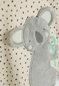Aplicación de peluche de koala gris sobre una tela beige con lunares negros; presenta orejas texturizadas y sostiene una ramita verde.