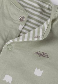 Hellgrünes Baumwollkleid mit weißen Kronenmuster, verziert mit silbernen Druckknöpfen und einem gestreiften Innenfutter. Verfügt über dunkelbraun gestickten Text.