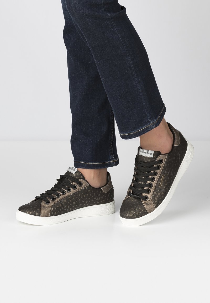 Brązowe sneakersy z gwiazdkowym wzorem, czarnymi sznurówkami i białą gumową podeszwą. Buty mają gładką fakturę i niski profil.