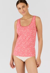 Top rosso senza maniche con un motivo floreale bianco, scollatura a barca e design aderente. Indossato con un pantalone di colore crema. Tessuto di morbida consistenza.