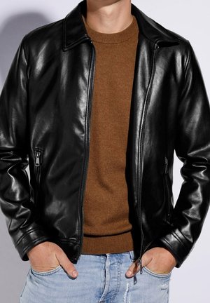 Man wearing black leather jacket over brown sweater with hands in pockets of light blue distressed jeans against plain background.