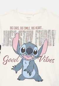 Personaje de dibujos animados Stitch con orejas grandes y sonrisa, el texto dice "Grandes orejas, gran sonrisa, gran corazón... Solo mantente divertido, Buenas Vibras" en tejido blanco.