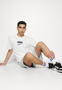 Biele bavlnené tričko s čiernym logom "PUMA", v kombinácii so svetlo šedými šortkami a čiernymi športovými topánkami, doplnené o biele a fialové pruhované ponožky.