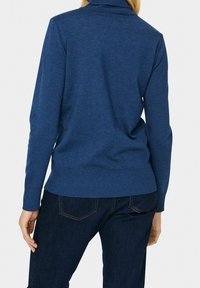 Niebieski sweter dzianinowy z wysokim kołnierzem, ściągaczami przy mankietach i dolnej krawędzi. Materiał wydaje się miękki i teksturowany. Noszony z ciemnymi dżinsami, prezentując casualowy wygląd.