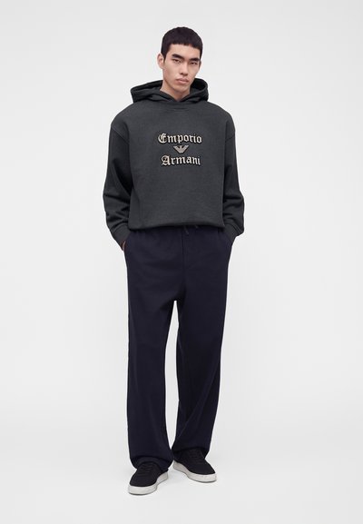 Jeune homme debout les mains dans les poches, portant un sweat à capuche foncé avec le logo Emporio Armani, un pantalon ample foncé et des baskets noires sur fond blanc.
