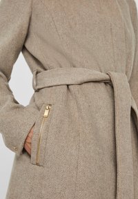 Beige ullkappa med en strukturerad herringbone-mönster, midja med bälte och en dragkedjeficka med detaljer i guld.