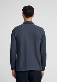 Mörkblå l ångärmad polo med krage, med en texturerad tyg och rak fåll, sedd bakifrån.