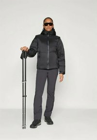 Črna izolirana jakna s kapuco, z zadrgo, v kombinaciji s temnimi hlačami in črnimi pohodniškimi čevlji. Drži črne pohodniške palice.