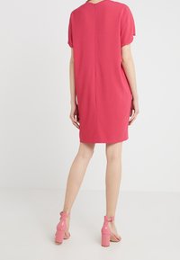 Kurzes pinkes Kleid mit kurzen Ärmeln, runder Halslinie und Reißverschluss im Rücken. Glattes Material mit lockerer Passform. Ein Paar pinke High Heels sind sichtbar.