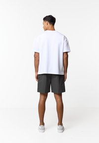 Άνδρας με την πλάτη, φοράει απλό λευκό κοντομάνικο μπλουζάκι, σκούρα γκρι σορτς μέχρι το γόνατο και λευκά αθλητικά παπούτσια, στέκεται σε λευκό φόντο.