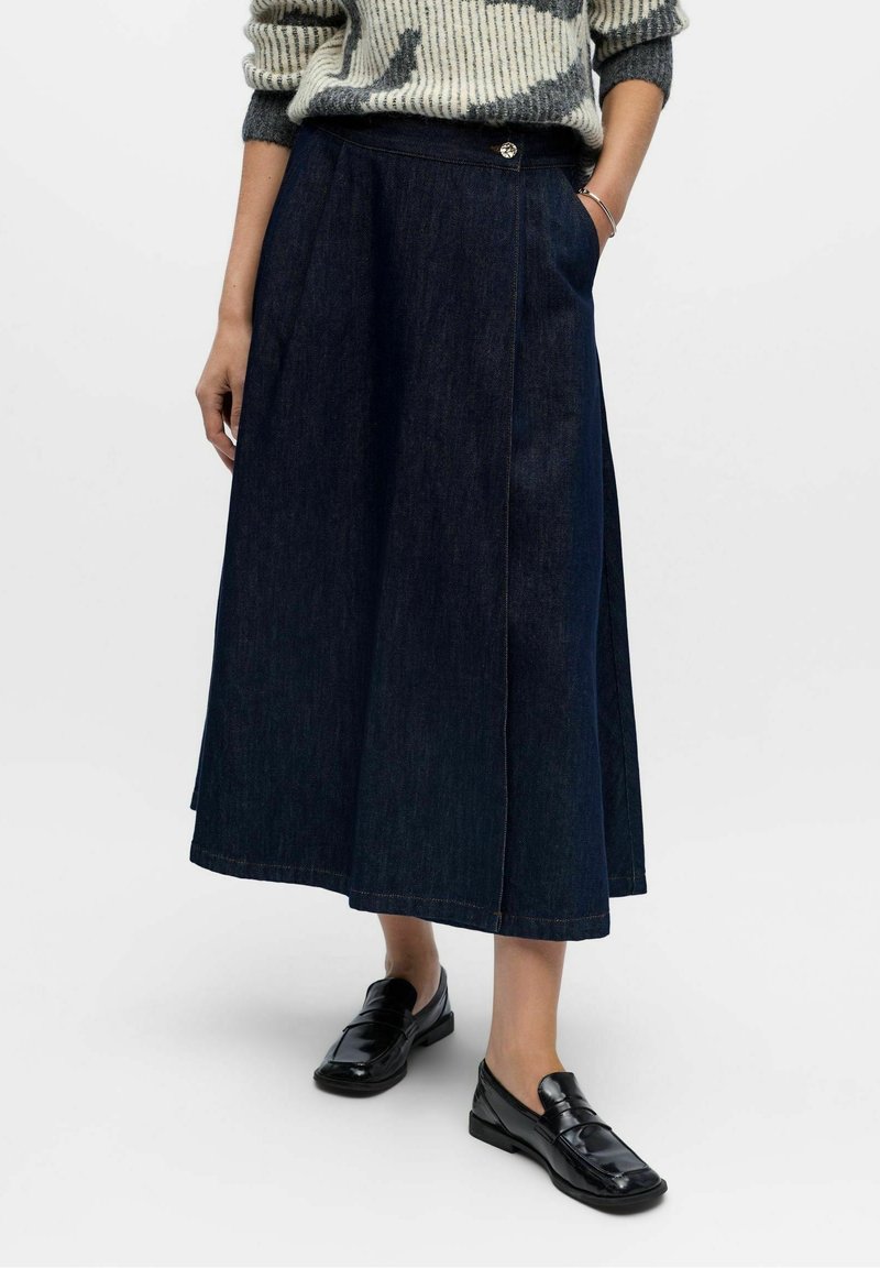 Personne portant une jupe midi en denim bleu foncé, des mocassins noirs et un pull crème et gris à motifs, debout devant un fond uni.