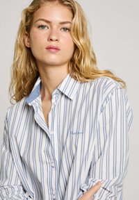 Chemise à boutons rayée bleue et blanche en tissu doux. Dotée d'un col bleu clair et d'un logo brodé sur la poitrine.