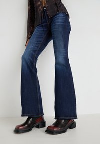 Jeans acampanados de un azul oscuro con una textura de denim suave, combinados con botas de tacón grueso en negro y burdeos y una blusa marrón texturizada.