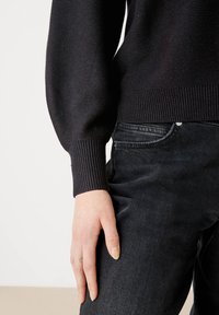 Zwarte, gestructureerde gebreide trui met ribbelmanchetten, gedragen over donkere denim jeans. De jeans hebben een zichtbare zak en subtiele stiksels.