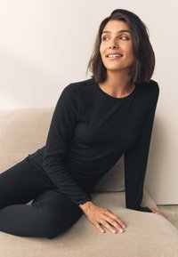 Top negro de manga larga con canalé y detalles de encaje en los puños, combinado con leggings negros a juego, sentado en un sofá de color claro.