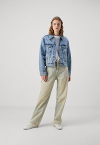 Ljusblå denimjacka med framfickor, bärs över en vit skjorta. Baggy ljusgröna byxor med slitna detaljer, i kombination med vita sneakers.