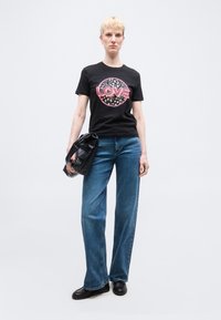 Άτομο με κοντά ξανθά μαλλιά φοράει μαύρο μπλουζάκι με το γράμμα "LOVE", μπλε τζιν, μαύρα παπούτσια και κρατάει μια μαύρη τσάντα χειρός με υφή.