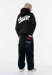 Schwarzer Kapuzenpullover mit weißem "Swd."-Logo auf dem Rücken, kombiniert mit lockeren schwarzen Jeans mit roten und weißen Akzenten sowie mehreren Taschen.