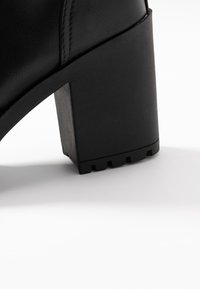 Mustaan korkokengät, jotka ovat neliömäiset, valmistettu teksturoidusta synteettisestä materiaalista. Varustettu tukeva lohkokorko ja uritettu pohja pidon parantamiseksi.