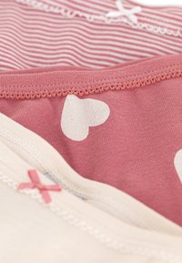 Três peças de vestuário sobrepostas: um top listrado rosa, uma peça rosa sólida com corações brancos e uma parte inferior creme com um detalhe de laço rosa.