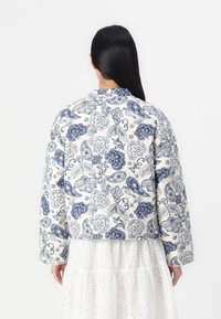 Pikowana kurtka w kwiatowy wzór w kolorach niebieskim i białym, z okrągłym dekoltem i szerokimi rękawami, zestawiona z fakturowaną białą spódnicą.