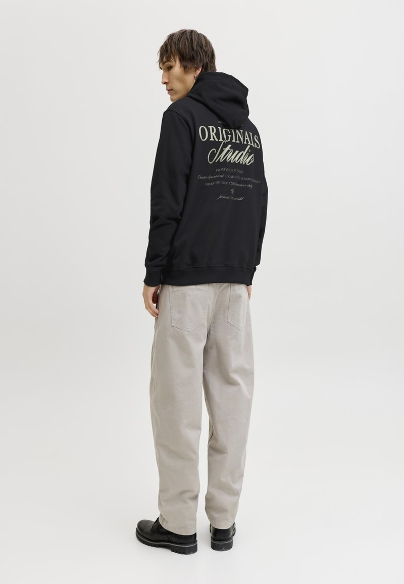 Homme portant un sweat à capuche noir avec le texte « Originals Studio » dans le dos, un pantalon beige et des chaussures noires, debout sur un fond clair uni.