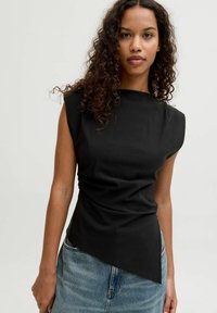 Sort ærmeløs top med asymmetrisk kant, lavet af glat stof. Har en høj halsudskæring og minimalistisk design. Kombineret med denim.