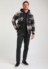 Gequilted geruite jas in blauw, bruin en groen, gecombineerd met een donkergroene hoodie en gestructureerde grijze broek, aangevuld met zwarte sneakers.
